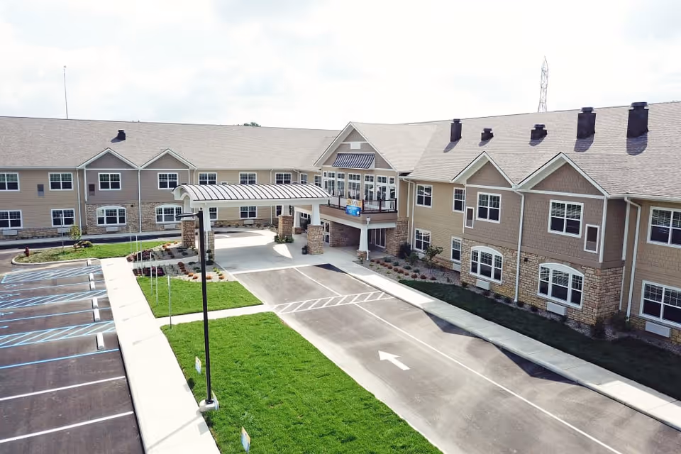 Exterior view of a senior living facility named The Glen, showing a two-story building with multiple windows, a covered entrance with stone pillars, a driveway with a white arrow, and a parking lot with marked spaces and green lawns.