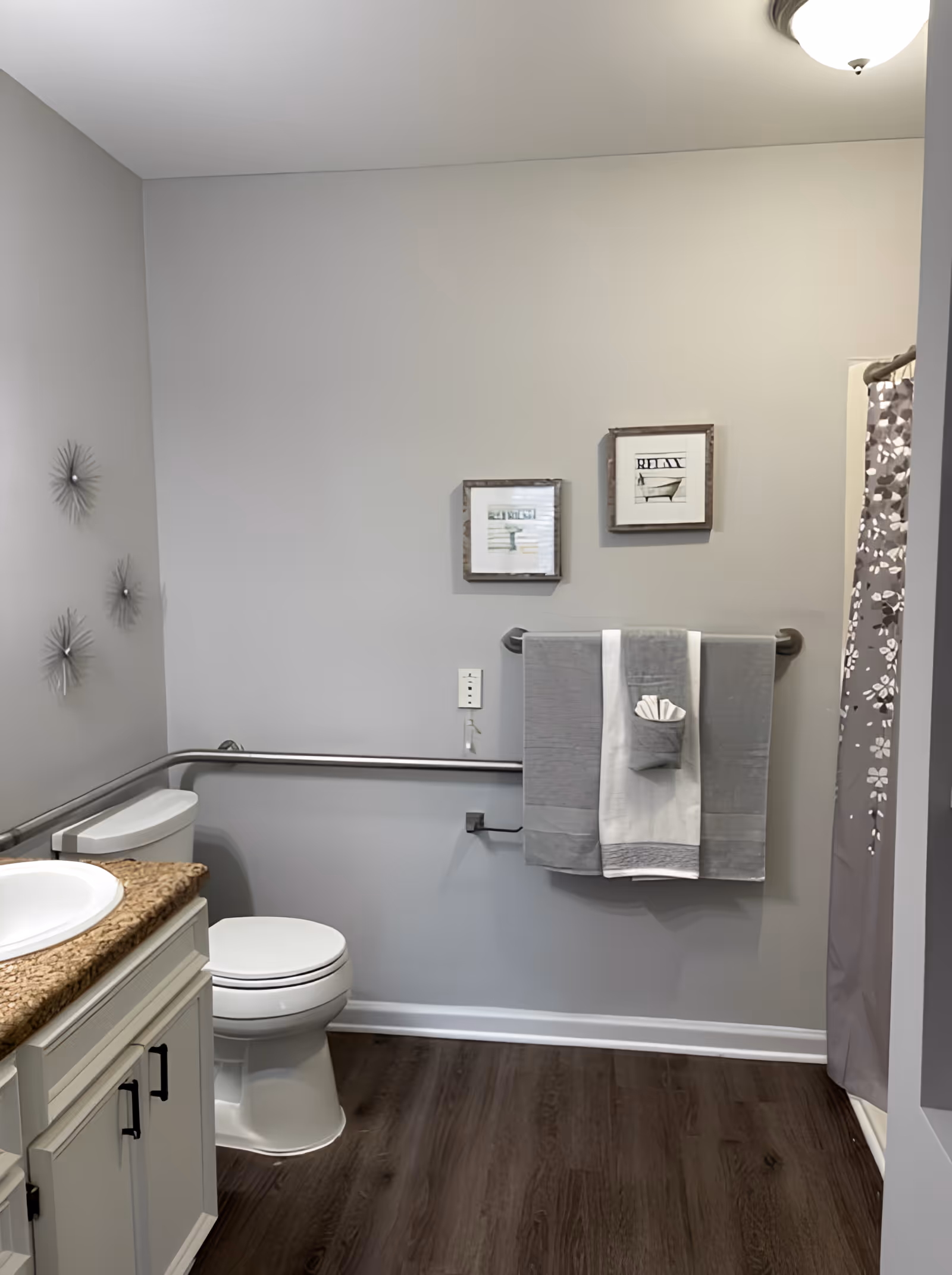A clean and modern bathroom with a white toilet, a granite countertop with a white sink, and a shower with a gray floral curtain. The walls are light gray with two framed pictures above a towel rack holding gray and white towels. There are decorative metal wall hangings on the left wall and a wooden floor.
