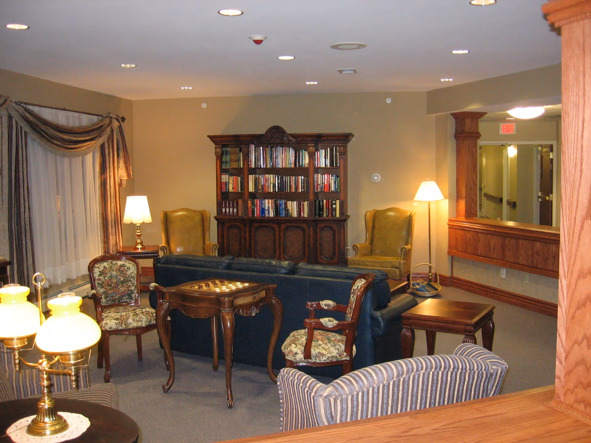 A cozy living room area with a dark blue sofa, two floral upholstered chairs, two yellow armchairs, a wooden bookshelf filled with books, a small wooden table with a chessboard, and several lamps providing warm lighting. The room has beige walls, carpeted floor, and a window with draped curtains.