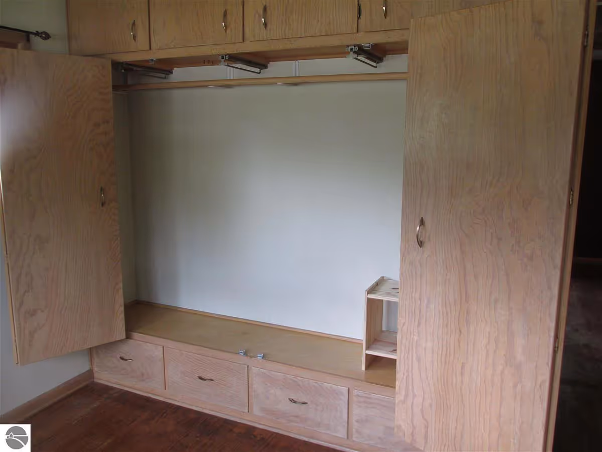 An empty wooden built-in wardrobe with open cabinet doors, three drawers at the bottom, and additional storage compartments above. The wardrobe is set against a plain wall and has a small wooden shelf inside on the right side. The floor is wooden.