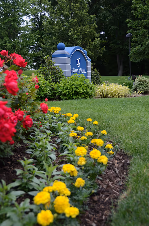 A landscaped garden area with vibrant red and yellow flowers in the foreground and a blue sign for The Grove at Valhalla Rehabilitation and Nursing Center in the background, surrounded by green bushes and trees.