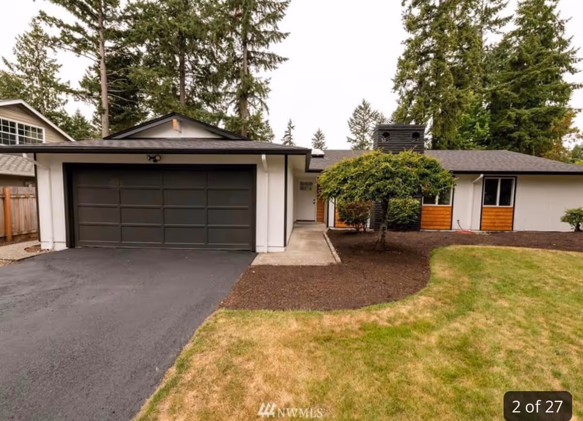 Front exterior view of a single-story house with a dark garage door, a small tree, and a lawn with patches of green and brown grass. The house has white walls with wooden accents and is surrounded by tall evergreen trees.