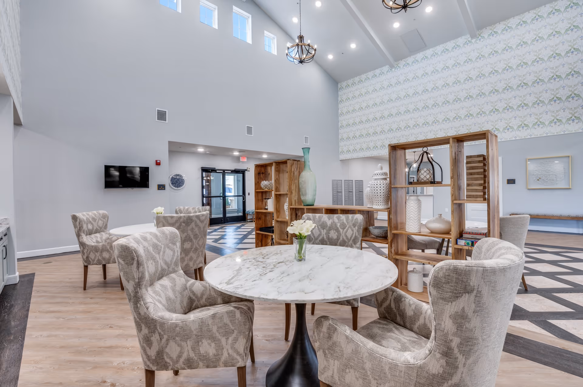 A bright and spacious common area with high ceilings and multiple windows near the ceiling. The room features round marble tables surrounded by patterned upholstered chairs. There are wooden shelving units with decorative vases and objects. The floor has a combination of light wood and a patterned dark wood design. A wall-mounted TV and a glass door entrance are visible in the background.