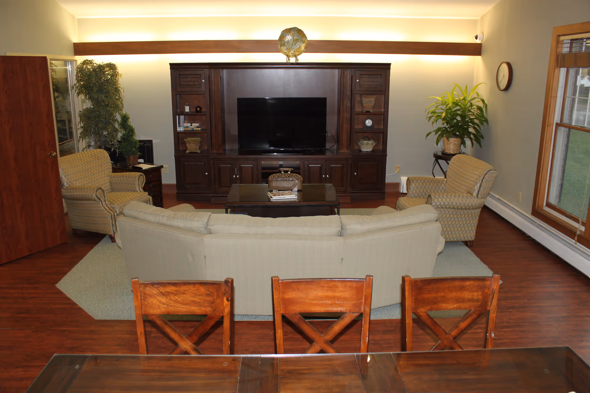 A cozy living room with a beige curved sofa, two patterned armchairs, a wooden coffee table, and a large dark wood entertainment center with a flat-screen TV. There are plants on either side of the entertainment center, a clock on the wall, and a window with wooden blinds letting in natural light. The floor is wooden with a light-colored area rug under the seating area.