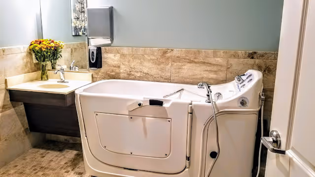 A bathroom with a walk-in bathtub featuring safety handles and controls, a sink with a faucet, a paper towel dispenser, a soap dispenser, and a vase of yellow and red flowers on the countertop. The walls are tiled halfway up with beige tiles and painted light blue above.