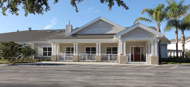 Single-story senior living building front with a covered columned entrance, windows, palm trees, and a parking area under a clear blue sky.