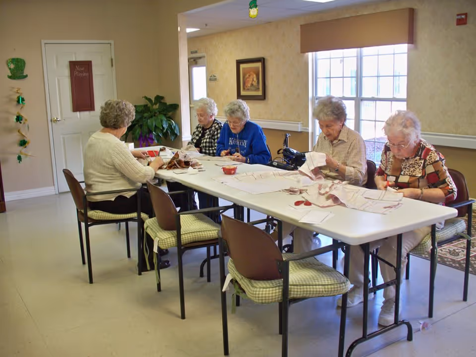 Five elderly women sitting around a rectangular table in a well-lit room, engaged in sewing or crafting activities. The room has beige walls, a window with a brown valance, a plant in the corner, and a door with a 'Now Playing' sign. Chairs with green checkered cushions surround the table.