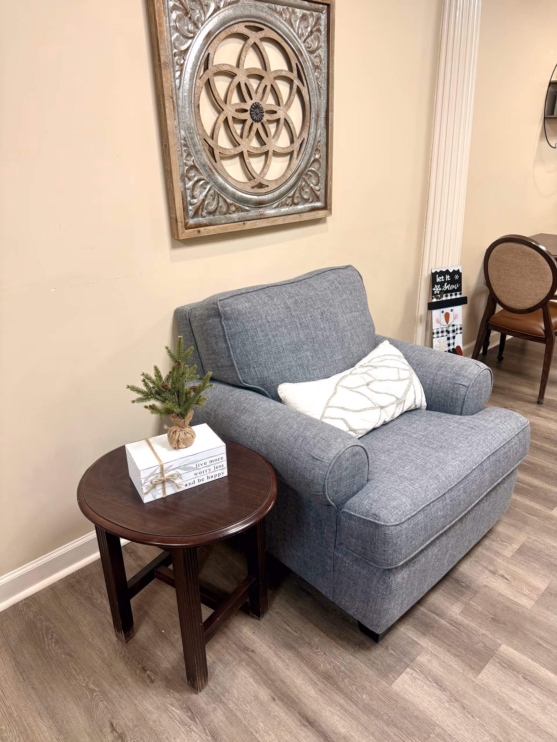 A cozy corner with a gray upholstered armchair featuring a white decorative pillow. Next to the chair is a round wooden side table with a small potted evergreen plant and a stack of books tied with string. Above the chair hangs a decorative wooden wall art piece with an intricate circular design. The floor is wood laminate, and part of a dining chair and table is visible in the background.