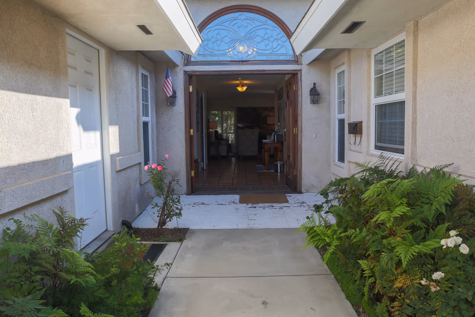 View of the entrance to a senior care facility with an open wooden door leading inside. The exterior walls are beige with windows on either side of the door. There are plants and flowers along the walkway leading to the entrance, and an American flag is mounted on the left wall near the door.