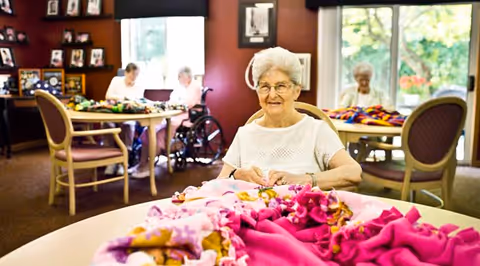 An elderly woman with white hair and glasses is sitting at a round table working on a colorful fabric project. In the background, three other elderly women are also seated at tables, engaged in similar activities. The room has large windows letting in natural light and is decorated with framed pictures on the walls.