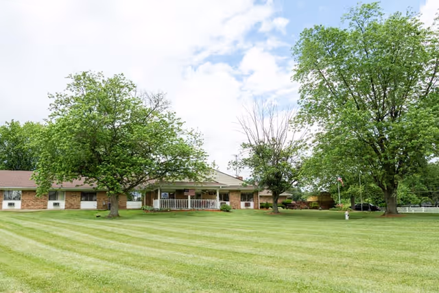 A single-story brick building with a covered porch and white railing, surrounded by a large, well-maintained grassy lawn and several mature trees under a partly cloudy sky.