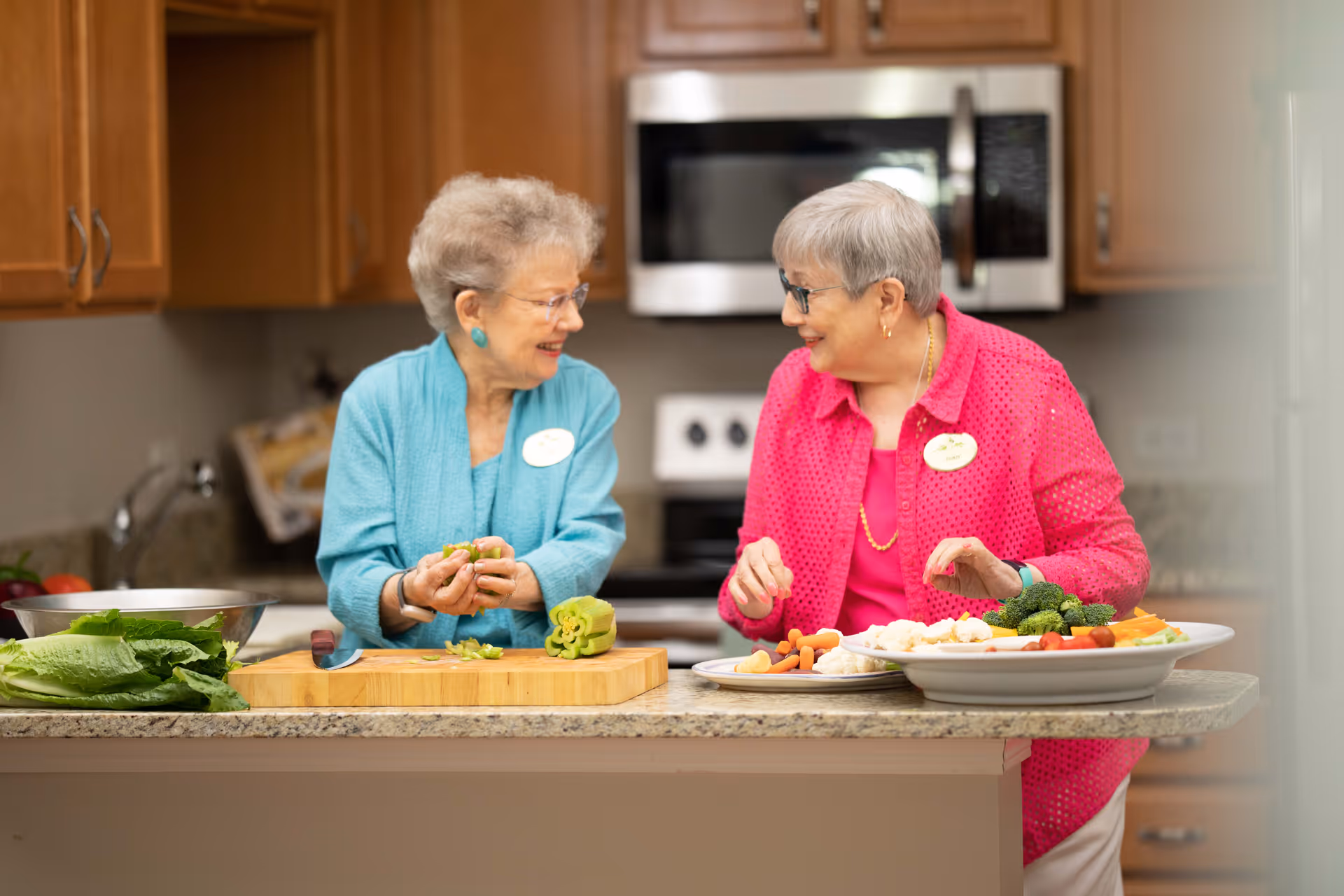Two elderly women smiling and preparing fresh vegetables together in a kitchen with wooden cabinets and a granite countertop. One woman is chopping green vegetables on a cutting board while the other arranges a plate of assorted vegetables including broccoli, carrots, and cauliflower.
