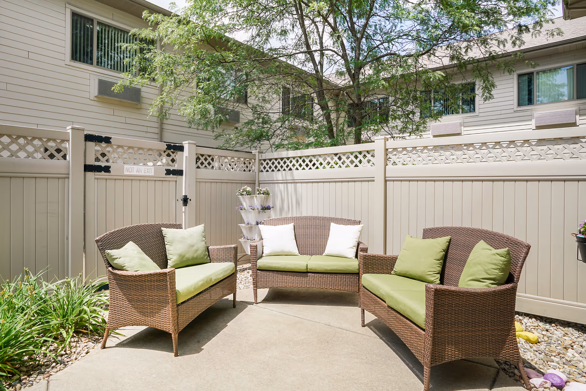 Sunny fenced courtyard patio with three wicker loveseats with green cushions arranged around a concrete area.