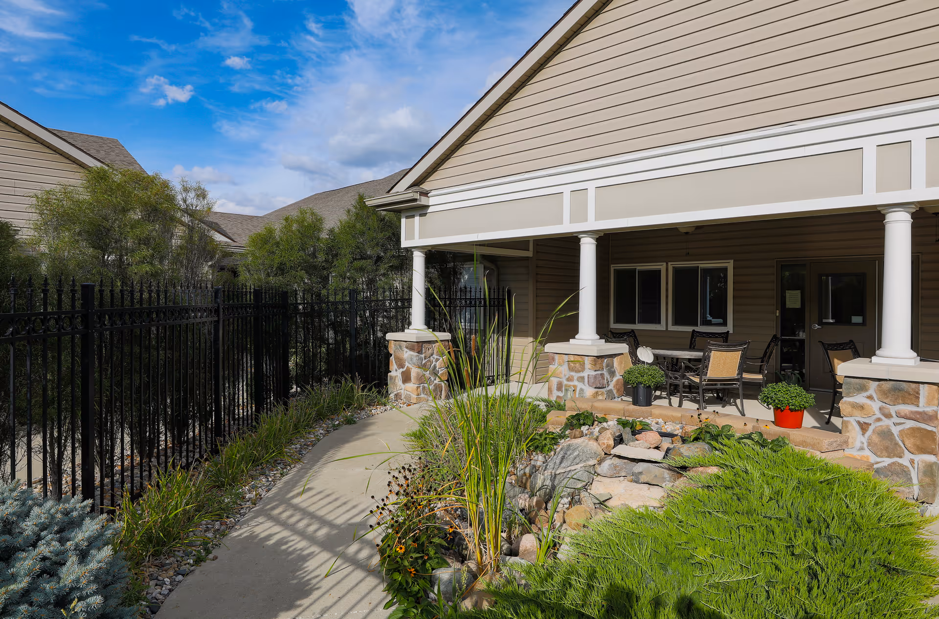 Outdoor patio area at Homestead Assisted Living & Memory Care of Creston featuring a covered seating area with a table and chairs, surrounded by greenery, plants, and a stone water feature. The patio is adjacent to the building with beige siding and white columns, under a partly cloudy blue sky.