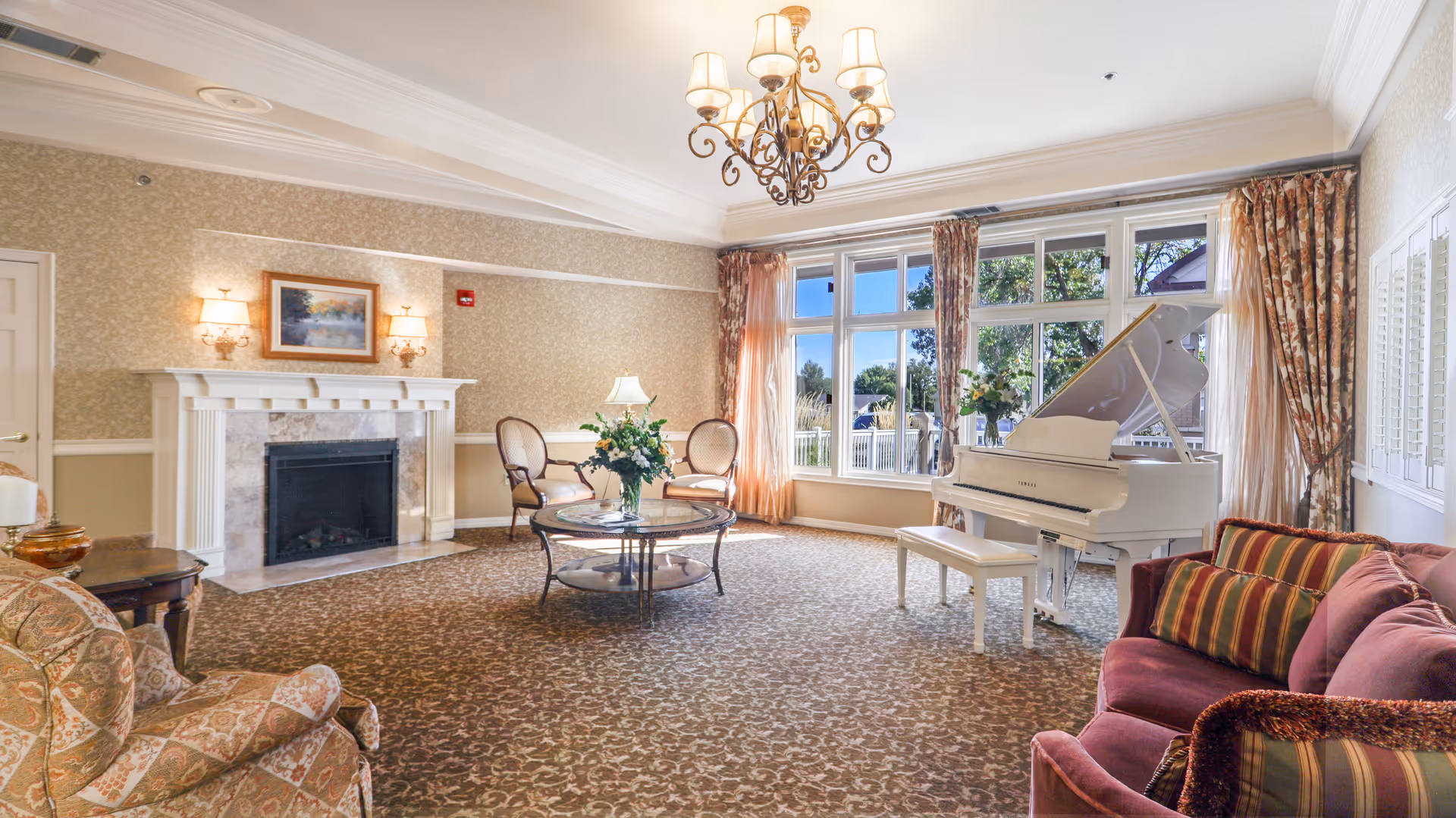 A spacious and elegant living room with a white grand piano near large windows with floral curtains. The room features a patterned carpet, a fireplace with a painting above it, a chandelier, a round glass coffee table with a flower arrangement, and comfortable upholstered chairs and a sofa.