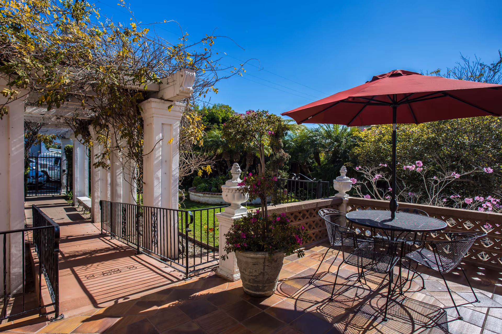 Outdoor patio area with a round metal table and four chairs under a large red umbrella. The patio is surrounded by a low decorative wall with potted plants and flowering bushes. A white pergola covered with vines is visible on the left side, leading to a gated entrance. The sky is clear and blue.