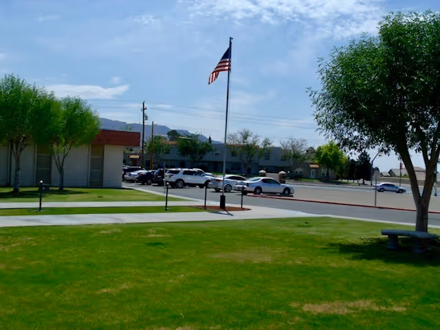 Outdoor view of Franklin Heights Nursing and Rehab Center showing a green lawn with trees, a flagpole with an American flag, a parking lot with several cars, and parts of the building under a partly cloudy sky.