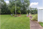 A grassy outdoor area with a concrete pathway leading through it. There are trees and bushes along the edges, a lamppost near the center, and a small covered structure in the background. The sky is partly cloudy.