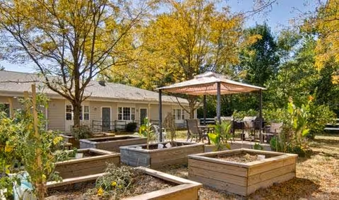 Outdoor garden area with raised wooden planter boxes, a patio umbrella shading a table with chairs, surrounded by trees and greenery, adjacent to a single-story building.