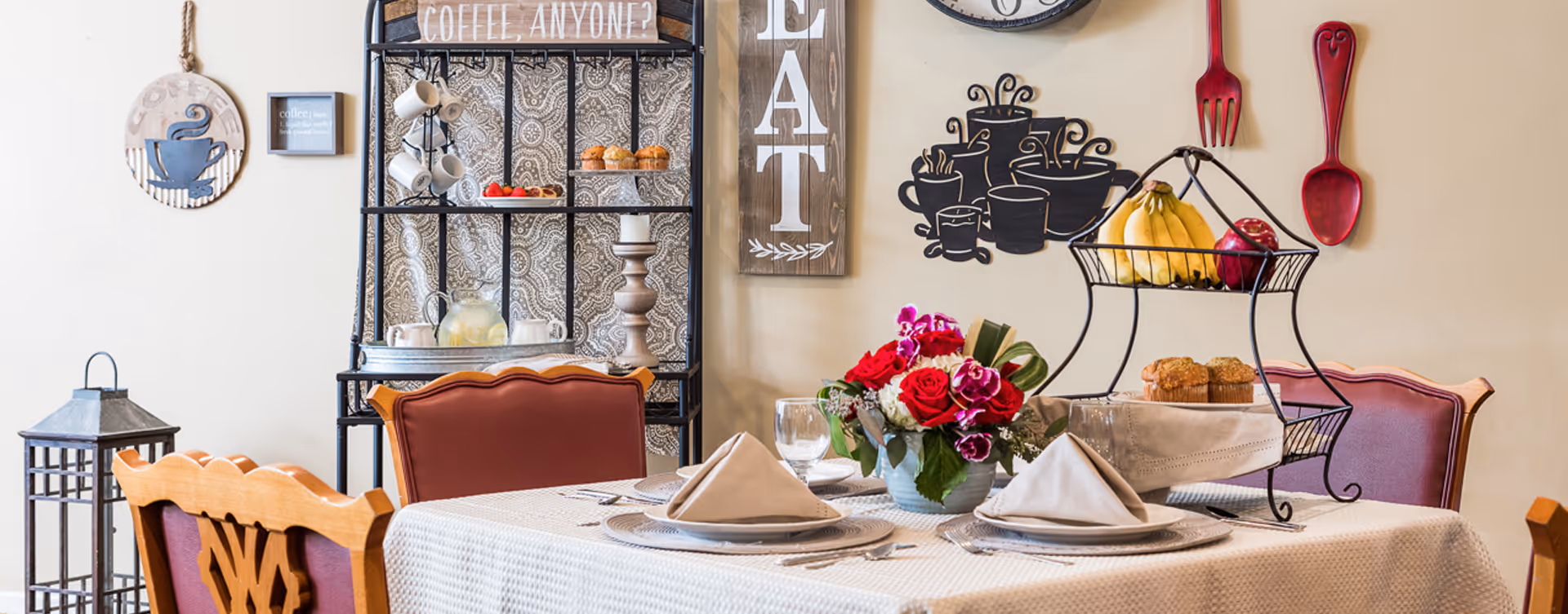 A dining table set for four with beige napkins folded on plates, a centerpiece of red and white flowers, and a two-tiered fruit basket with bananas and an apple. Behind the table is a black metal shelf with mugs, muffins, and a pitcher of lemonade. The wall is decorated with wooden signs, including one that says 'COFFEE, ANYONE?' and another that says 'EAT', along with large red wooden fork and spoon decorations.