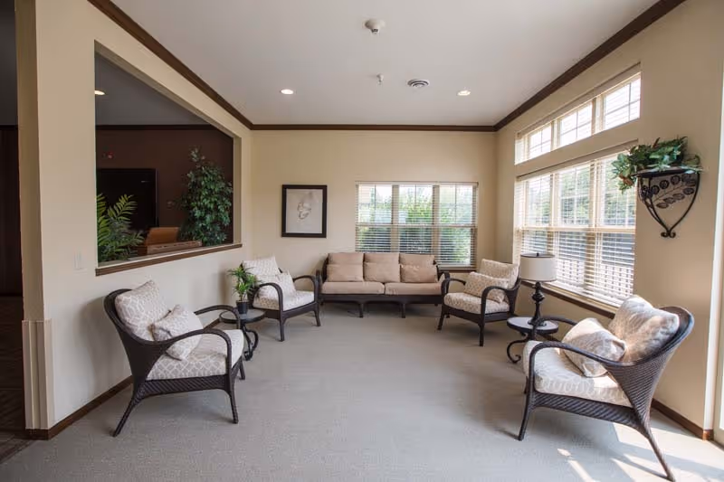 A bright and spacious living room with beige walls and carpeted floor. The room features a beige sofa and four cushioned armchairs arranged around small side tables. Large windows with blinds let in natural light, and there is a decorative wall-mounted plant holder with greenery on the right wall. A framed artwork hangs on the left wall, and a large interior window opens to another room with plants visible.