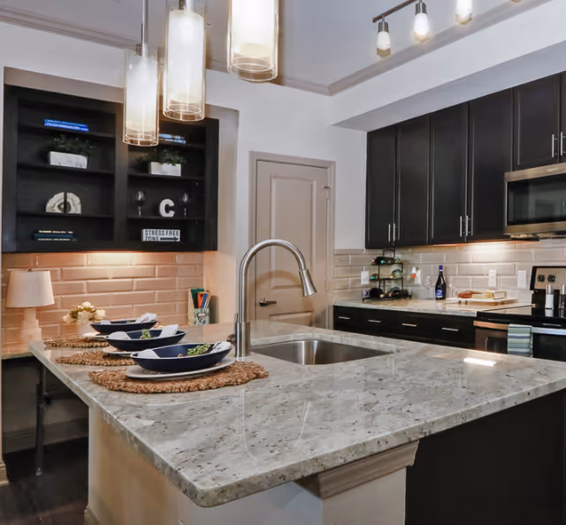 Modern kitchen with a large granite island countertop featuring a built-in sink and a sleek faucet. The island is set with three place settings including blue bowls and woven placemats. The kitchen has dark wood cabinets, stainless steel appliances including a microwave and stove, and a beige subway tile backsplash. Pendant lights hang above the island, and there are decorative items on the shelves and countertop.