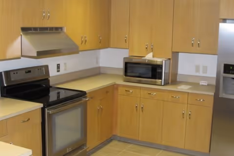 A kitchen area with light wood cabinets, a stainless steel electric stove with oven, a stainless steel range hood, a microwave on the countertop, and a stainless steel refrigerator with a water and ice dispenser. The countertops are beige, and the floor is tiled.