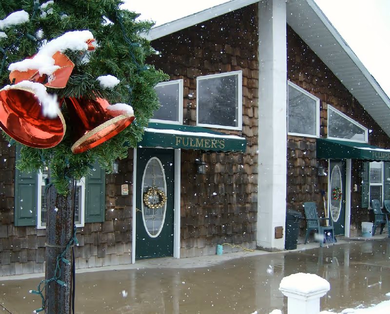 Exterior view of Fulmer's Personal Care Home building with brown wooden shingles, green doors with oval windows decorated with wreaths, green awnings with the name 'FULMER'S', snow falling, and a close-up of a snow-covered Christmas wreath with red bells on a pole in the foreground.