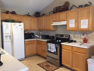 A kitchen with wooden cabinets, a white refrigerator, a stainless steel electric stove with a range hood, a microwave, a toaster, and various kitchen items on the countertops. There is a small rug in front of the stove and decorative items on top of the cabinets.