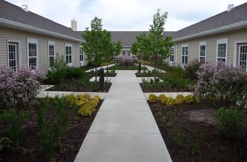 Paved walkway through a landscaped courtyard with shrubs and small trees between single-story senior living buildings.