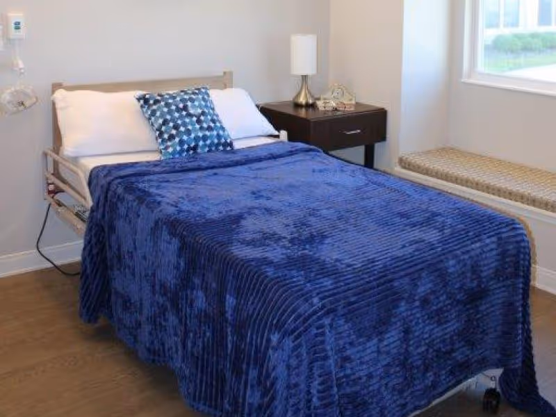 Single bed with a dark blue blanket in a small, neatly furnished bedroom with a nightstand, lamp, and window seat.