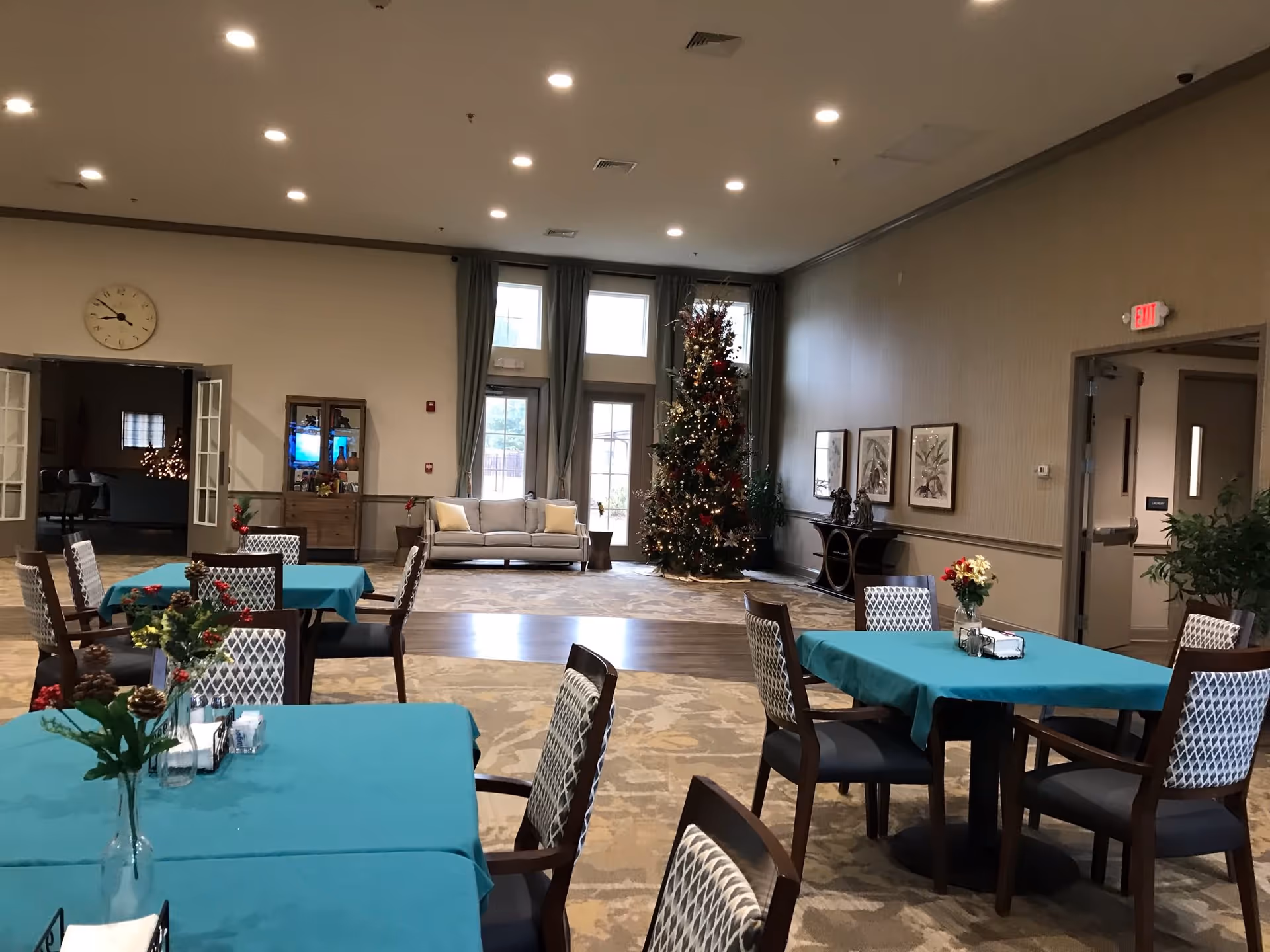 Interior view of a senior living facility common area decorated for the holidays with a Christmas tree in the corner. Several tables with teal tablecloths and chairs are arranged in the foreground, each table adorned with small floral centerpieces. A beige sofa is positioned near large windows with curtains, and framed artwork hangs on the walls. The room is well-lit with recessed ceiling lights.