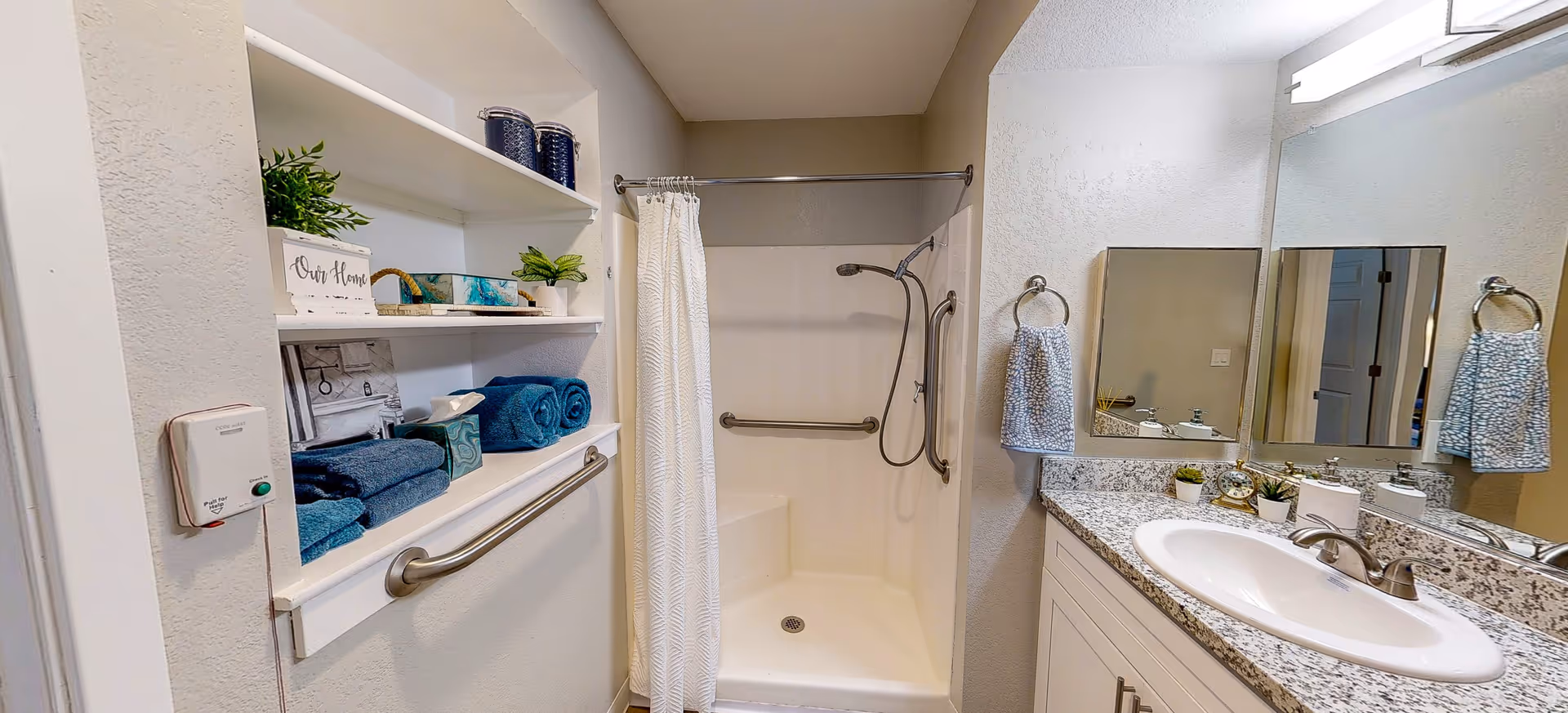 A bathroom with a walk-in shower featuring grab bars and a handheld showerhead. There is a white shower curtain, a granite countertop with a sink, a large mirror, and two towel rings with patterned towels. Shelves on the left hold folded blue towels, decorative plants, and storage containers. A pull cord emergency alert button is mounted on the wall near the shelves.