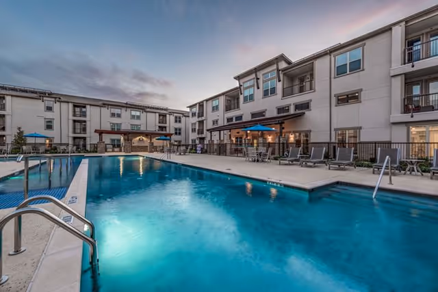 Outdoor swimming pool area at a senior living facility with lounge chairs, tables with umbrellas, and a multi-story building surrounding the pool under a twilight sky.