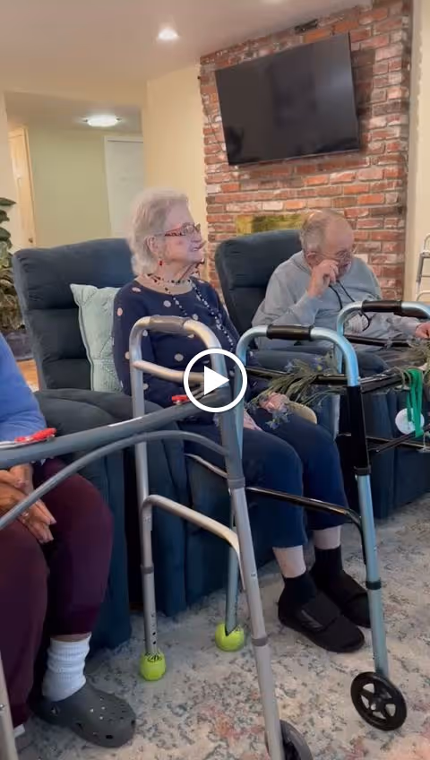 Elderly residents sit in recliners with walkers in a cozy living room featuring a brick fireplace and wall-mounted TV.