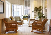 A bright sunroom with large windows featuring wicker furniture including chairs and a small table, decorated with potted plants and flowers, creating a cozy and inviting sitting area.