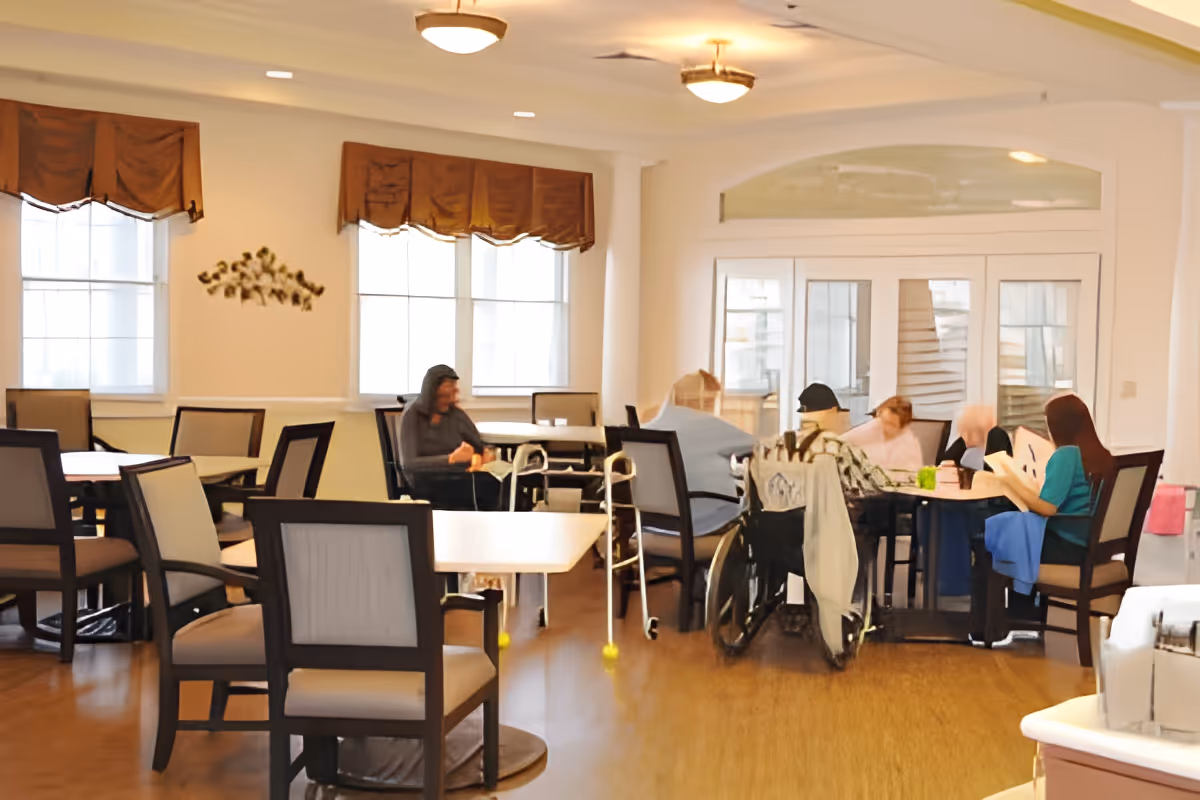 A bright dining/activity room with seniors and staff seated around tables, some using a wheelchair and walker.