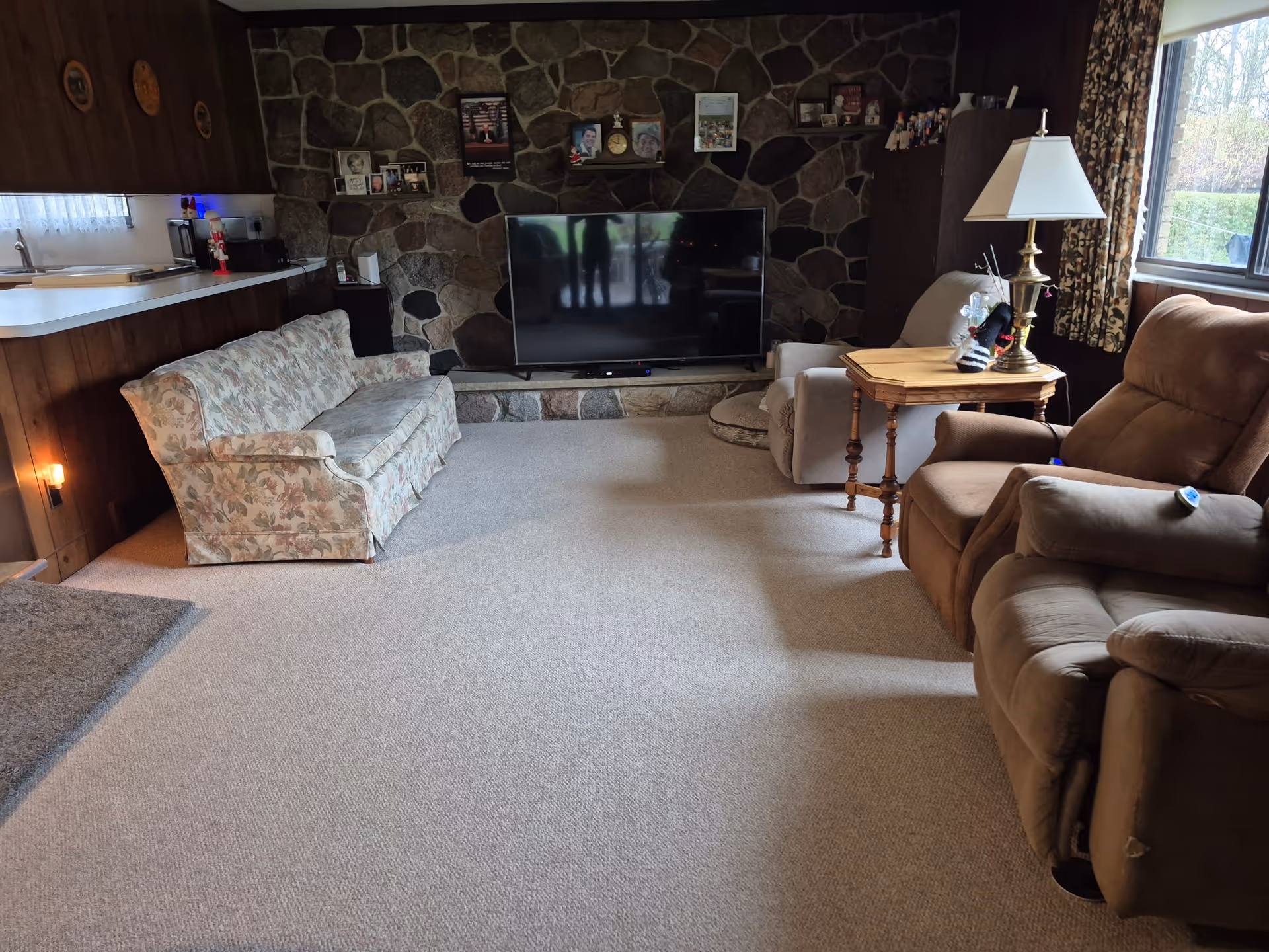 A cozy living room with a stone wall featuring a flat-screen TV mounted on a stone hearth. The room has a floral-patterned couch on the left, two recliner chairs on the right, and a wooden side table with a lamp and decorative items. There is a window with floral curtains letting in natural light, and a kitchen counter with a sink and microwave is partially visible on the left side.