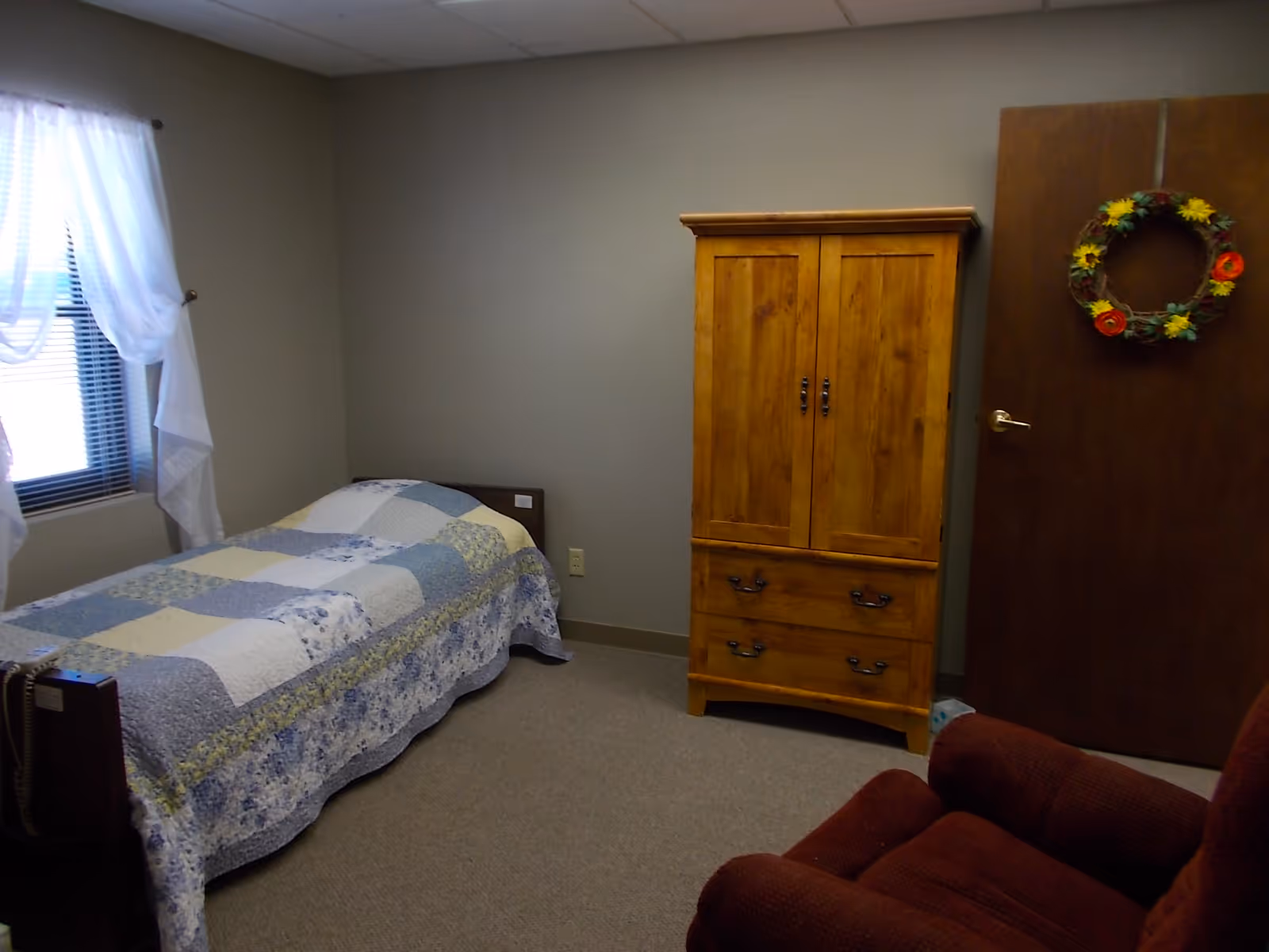 A senior living bedroom with a single bed, wooden armoire, and a wreath on the door.