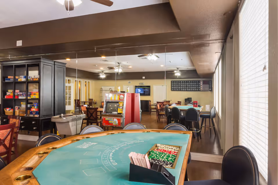 Activity room with a green felt poker table in the foreground, chairs, game shelves, and a slot machine reflected in a large mirror.