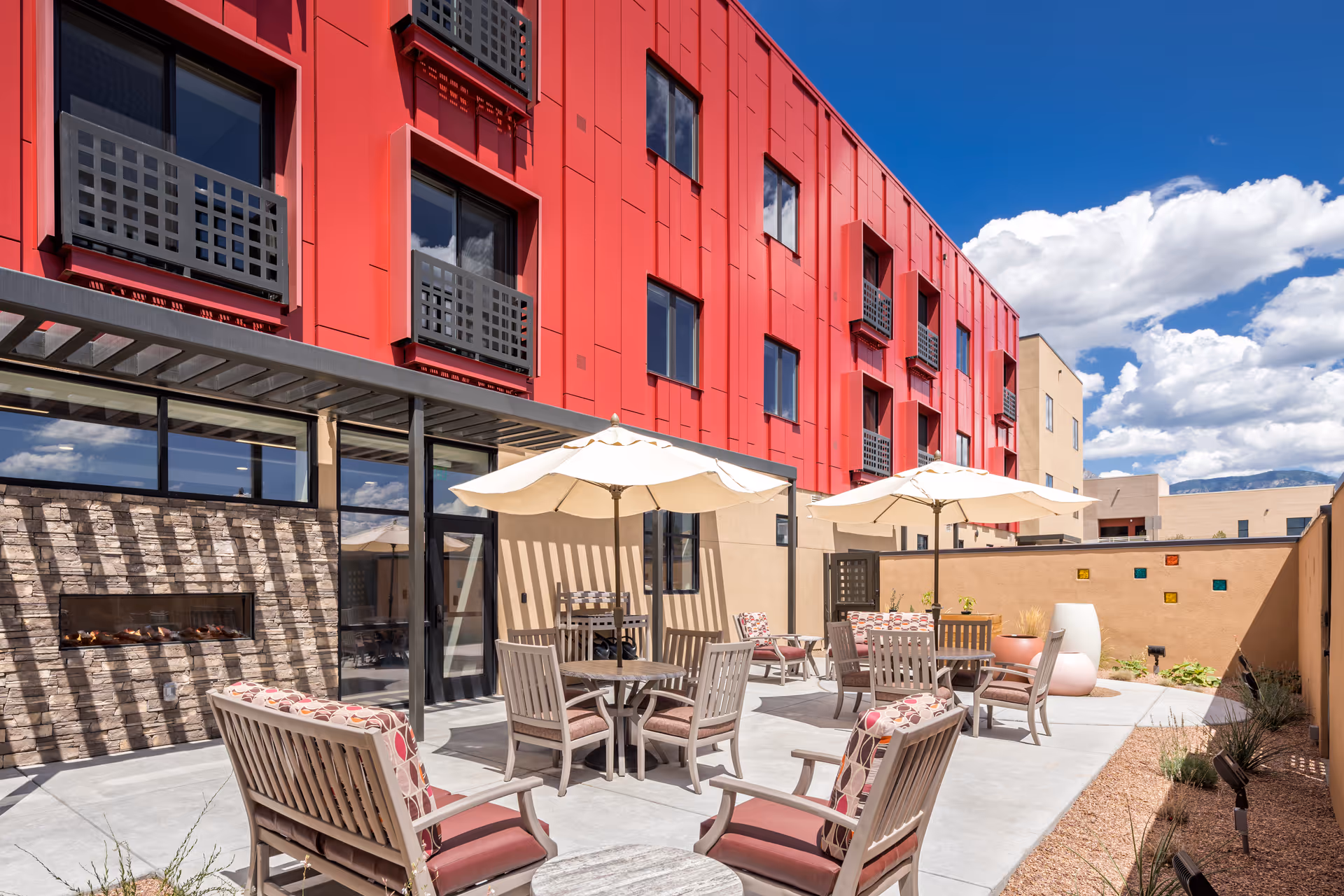 Outdoor patio area at Amaran Senior Living with several wooden chairs and tables under large white umbrellas. The patio is adjacent to a building with a red exterior and multiple windows. There is a stone wall with a built-in fireplace and a clear blue sky with some clouds overhead.