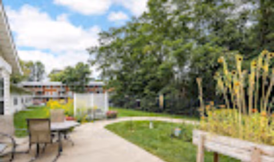 Outdoor courtyard with a patio table and chairs, a winding concrete path, garden beds, and trees.