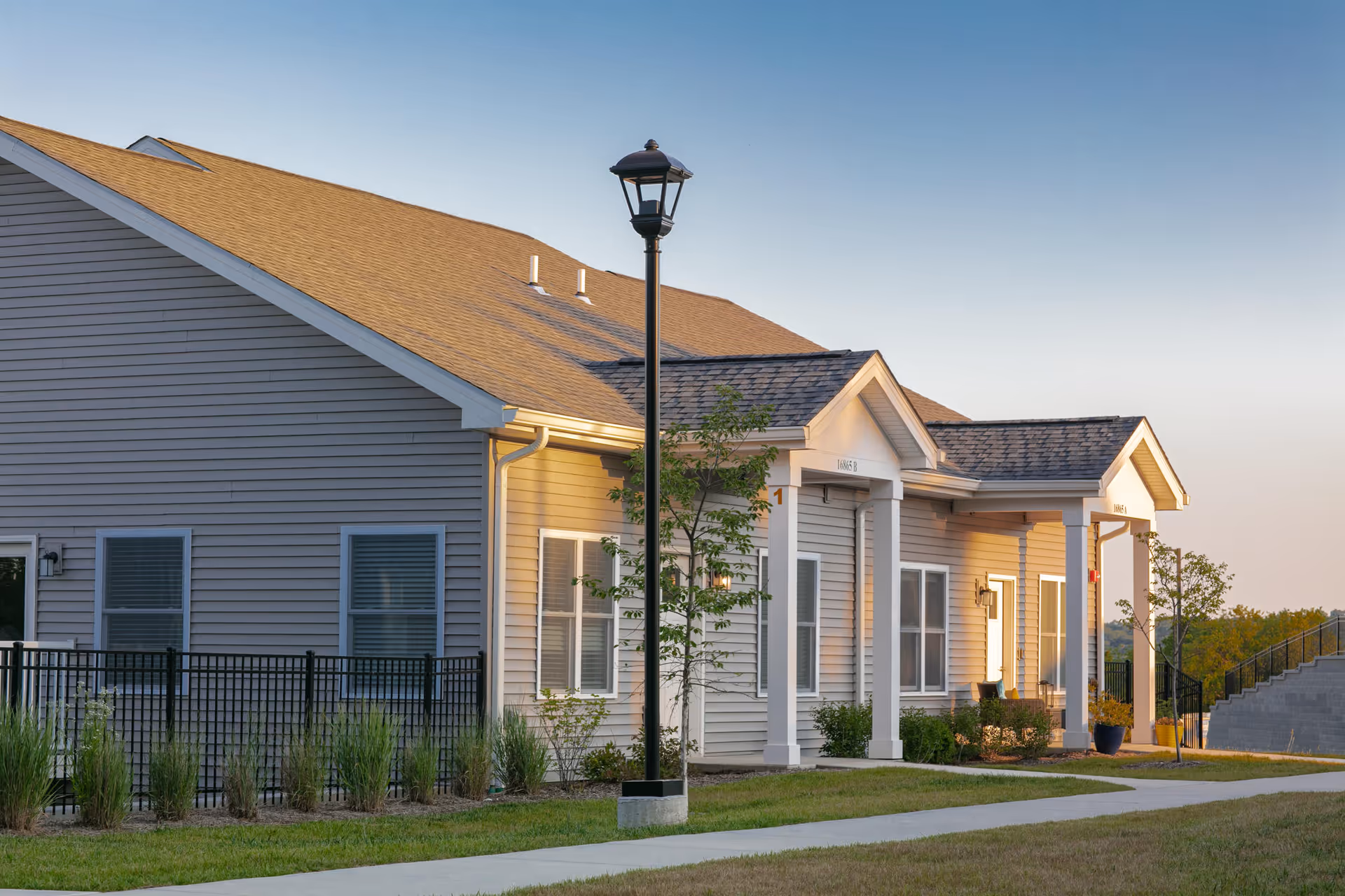 Exterior view of a single-story residential building with beige siding and a gabled roof, featuring white columns at the entrance, a black metal fence, a sidewalk, and a street lamp in the foreground during early evening.