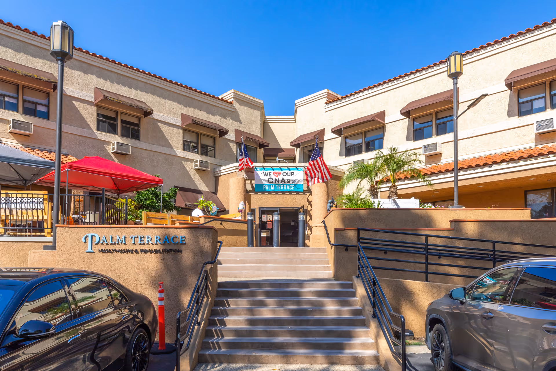 Front entrance of Palm Terrace Healthcare & Rehab Center with stairs, signage, American flags, a patio area, and parked cars.