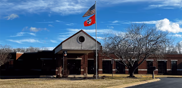 Front exterior view of a single-story brick building with a peaked entrance. Two flags, the American flag and the Tennessee state flag, are flying on a flagpole in front of the building. There is a leafless tree on the right side and a paved driveway leading up to the entrance under a partly cloudy blue sky.