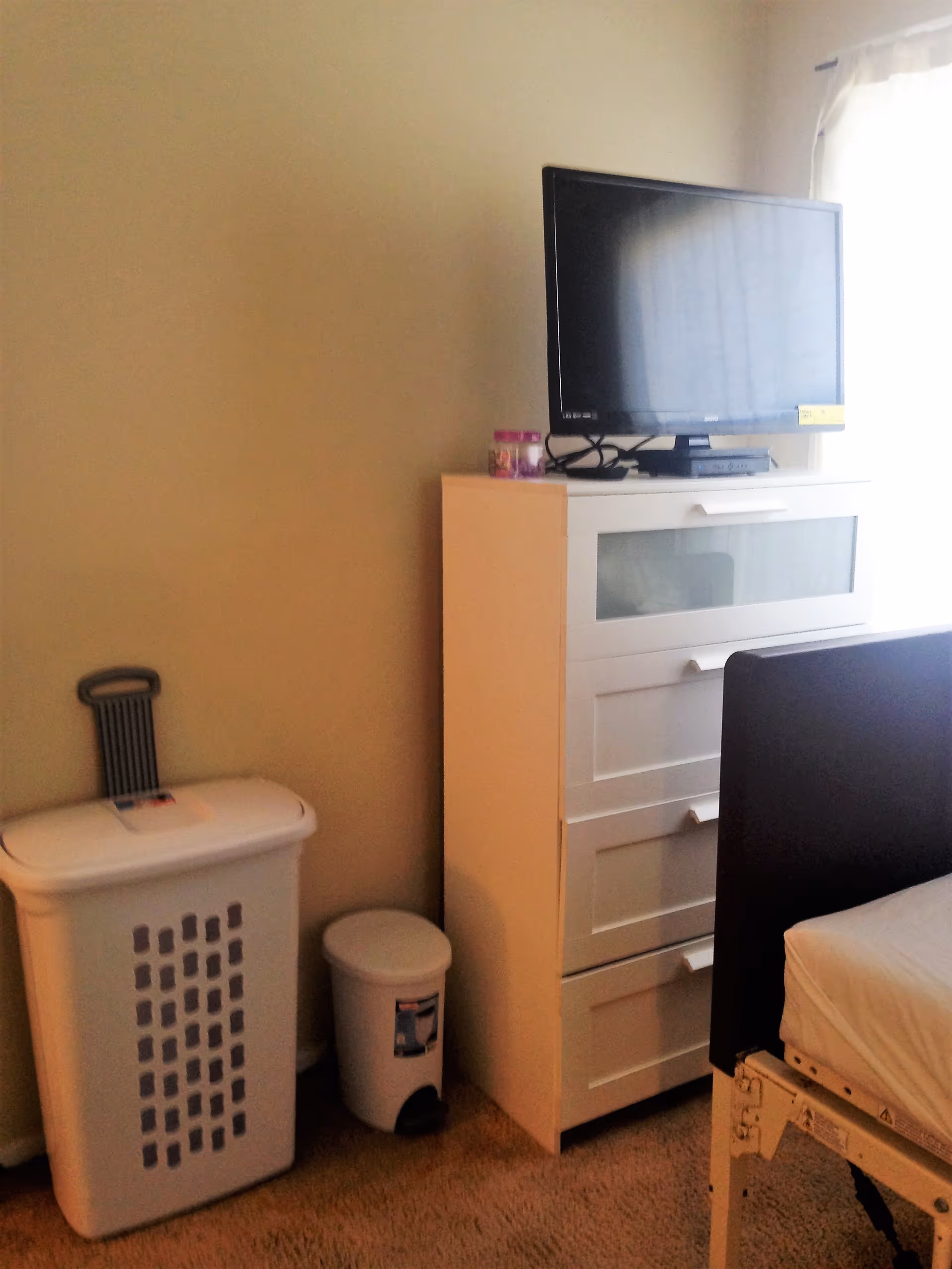 A corner of a bedroom featuring a white chest of drawers with a flat-screen TV on top, a white laundry basket with a handle, a small white trash bin, and part of a bed with a white mattress and dark headboard. Light filters in from a window with sheer curtains.