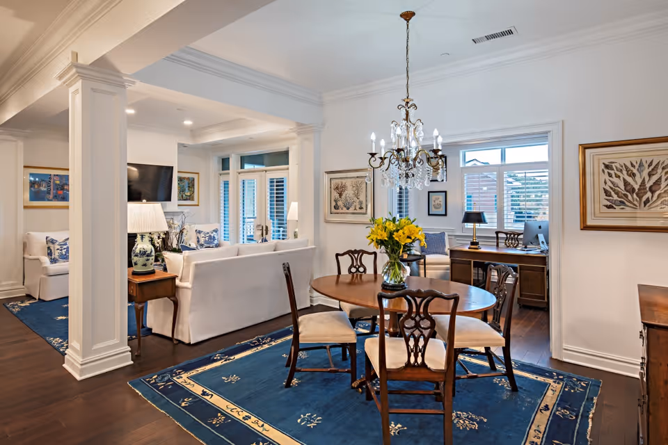A bright and elegant living and dining area featuring a round wooden dining table with four chairs on a blue patterned rug. A crystal chandelier hangs above the table. The living area has white sofas with blue and white cushions, a side table with a lamp, and a wall-mounted TV. Large windows with white shutters allow natural light to fill the room. The walls are adorned with framed artwork, and a wooden desk with a computer and lamp is visible in an adjacent room.
