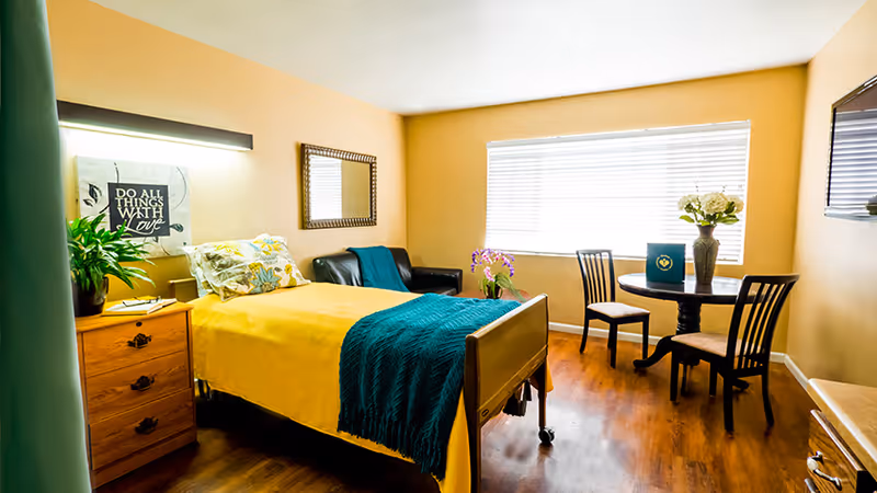 Sunny furnished bedroom with a single bed covered in yellow linens and a teal throw, a nightstand, armchair, small round dining table and large window.