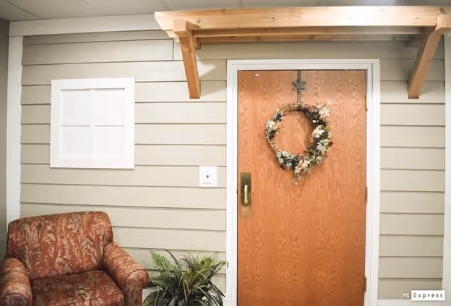 A cozy indoor corner featuring a wooden door decorated with a floral wreath, a patterned armchair, a potted plant, and light-colored horizontal wall paneling with a small white window.