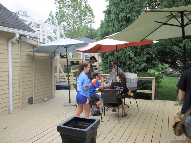 A group of people gathered on a wooden deck outdoors with large umbrellas providing shade. There are chairs and a table with a cooler on it, surrounded by trees and greenery.